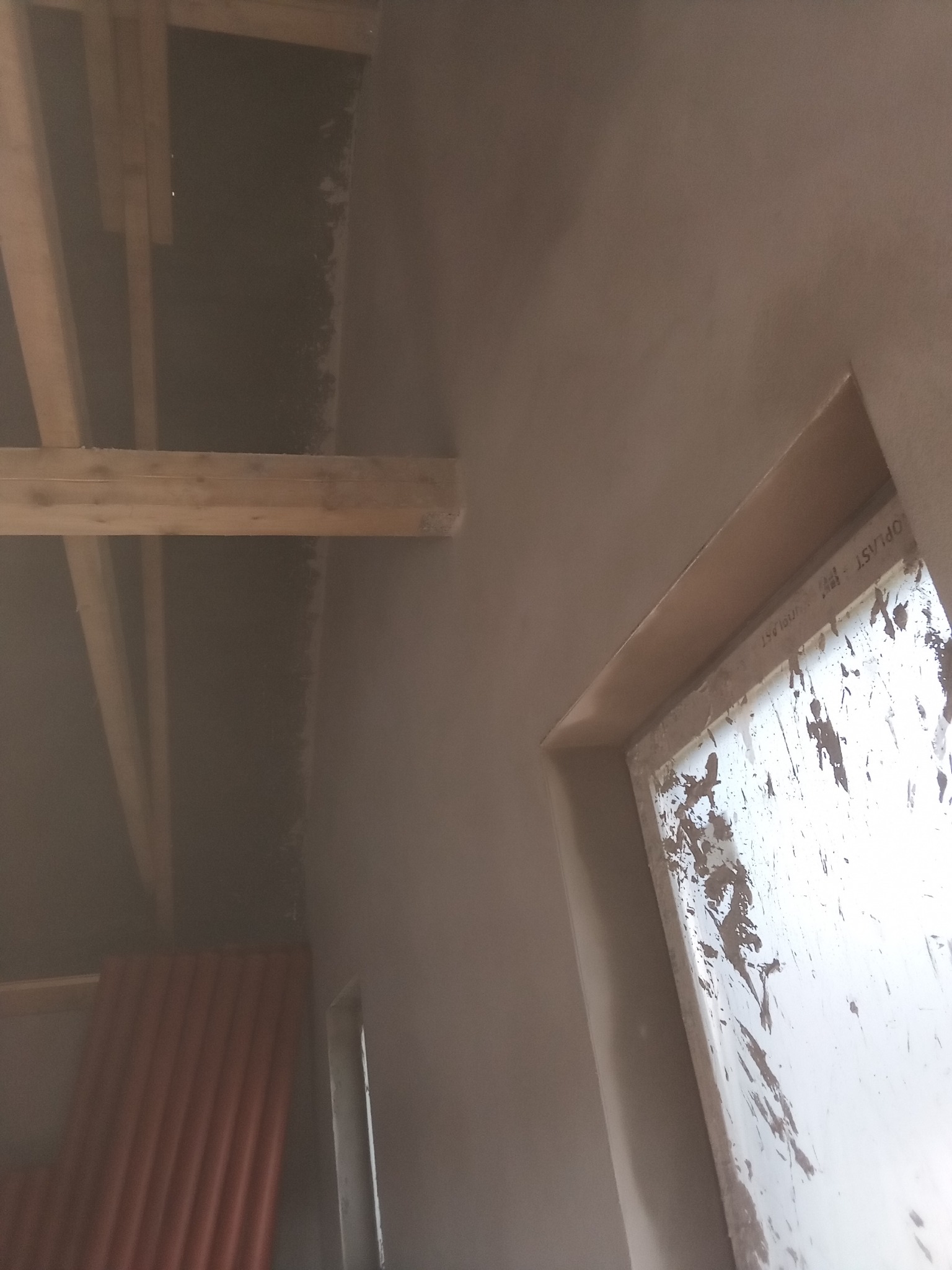 Świeżo nałożone tynki maszynowe na ścianach poddasza z widocznymi drewnianymi belkami stropowymi i oknem zabrudzonym zaprawą.