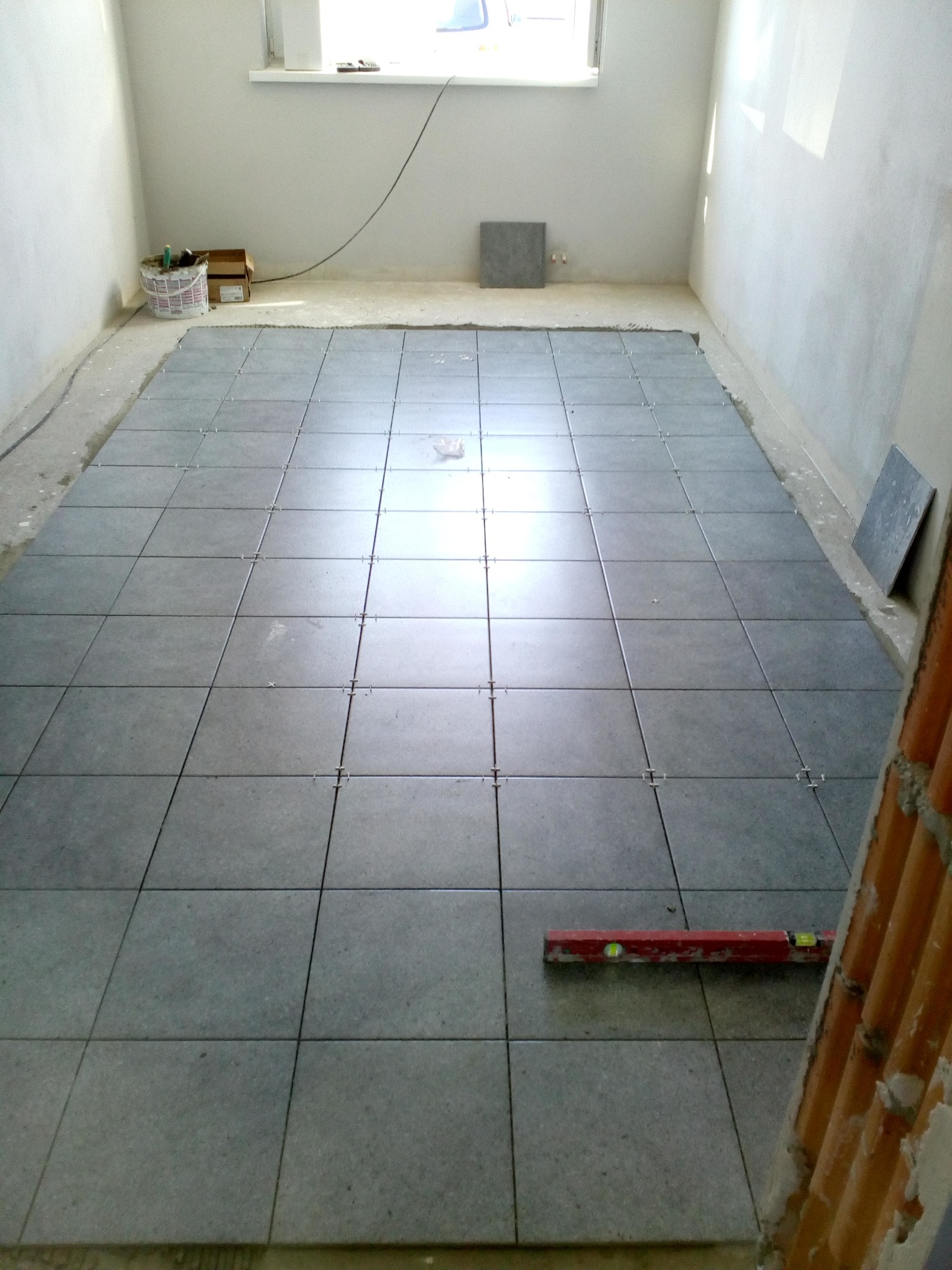 Szare płytki podłogowe ułożone z krzyżykami dystansowymi w trakcie remontu pomieszczenia, widoczna poziomica i wiadro z narzędziami.