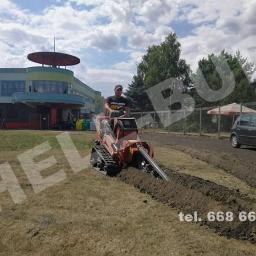 HELL-BUD Heleniak Adrian - Operator na minikoparce łańcuchowej wykopuje wąski rowek w ziemi na trawniku, w tle budynek z charakterystycznym okrągłym dachem i zaparkowany samochód osobowy.