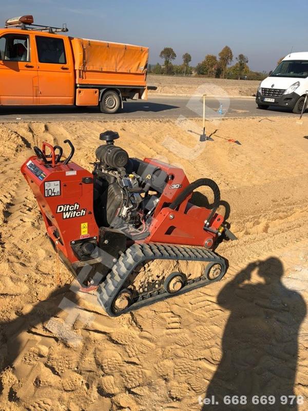 Pomarańczowa koparka łańcuchowa Ditch Witch 30 na piasku z widocznym numerem telefonu, w tle pomarańczowy samochód dostawczy i biały bus