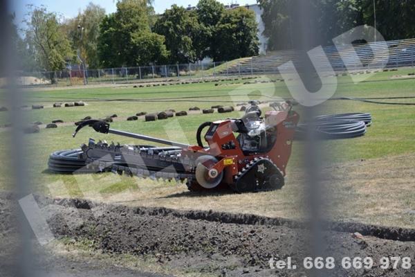Pomarańczowa maszyna do wykopów rowów na trawiastym terenie, z widocznymi rolkami darni i rurami w tle, częściowo zasłonięta przez siatkę ogrodzeniową.