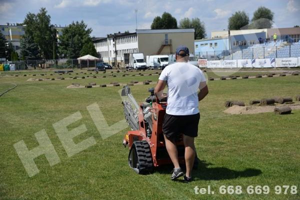 Operator prowadzi minikoparkę łańcuchową na murawie boiska, w tle budynki i trybuny stadionu, świeżo wycięte pasy darni ułożone w rzędach.