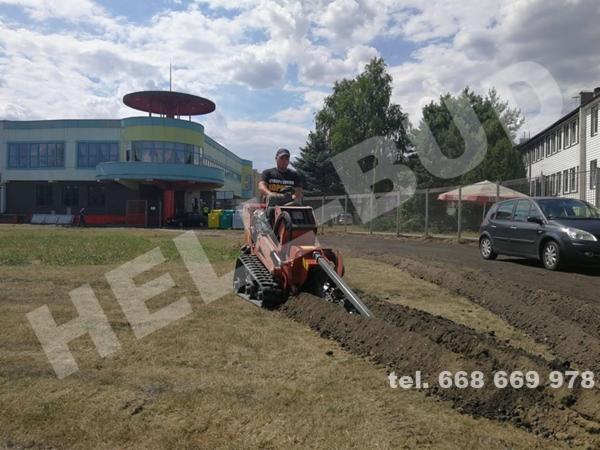 Operator na minikoparce łańcuchowej wykopuje wąski rowek w ziemi na trawniku, w tle budynek z charakterystycznym okrągłym dachem i zaparkowany samochód osobowy.