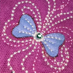 Zbliżenie na fioletową tkaninę z aplikacją w kształcie motyla z jasnoniebieskiego filcu, ozdobioną białym haftem i zielonym dżetem w centrum.