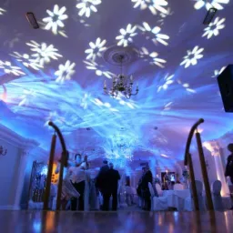 Sala bankietowa z oświetleniem dekoracyjnym: białe kwiaty wyświetlane na suficie, niebieskie światło wypełniające przestrzeń, żyrandol i kolumny w tle.