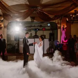 Para młoda tańczy pierwszy taniec w chmurze dymu na tle sceny z nagłośnieniem i oświetleniem, goście obserwują.