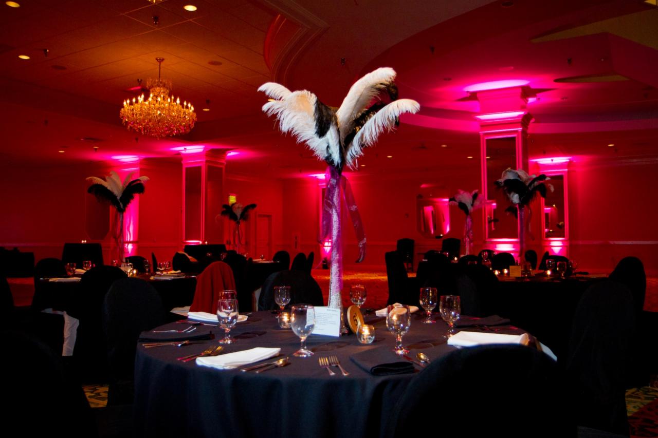 Uroczysta sala bankietowa z okrągłymi stołami nakrytymi czarnymi obrusami, białymi serwetkami i szklanymi naczyniami, oświetlona czerwonymi światłami i ozdobiona wysokimi wazonami z piórami.
