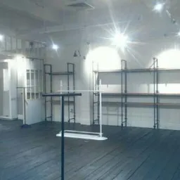 Półki metalowe na ścianie i wieszaki na ubrania w pustym, jasnym pomieszczeniu w Kołobrzegu, z drewnianą podłogą pomalowaną na ciemno i białymi ścianami, przygotowane do aranżacji sklepu.