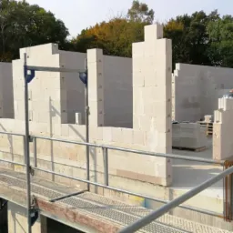 Wznoszenie ścian parteru z białych bloczków na placu budowy, widoczne rusztowanie i pracownik w niebieskim kasku.