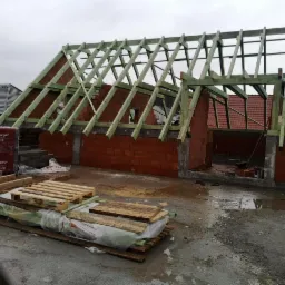 Wznoszenie ścian z czerwonej cegły i konstrukcji drewnianej dachu na budowie domu jednorodzinnego w pochmurny dzień. Na pierwszym planie palety z materiałami budowlanymi.