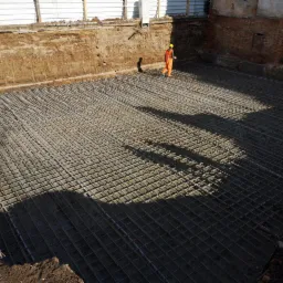 Zbrojenie fundamentów na placu budowy, widoczny pracownik w pomarańczowym kombinezonie i żółtym kasku, przygotowany wykop pod budynek.