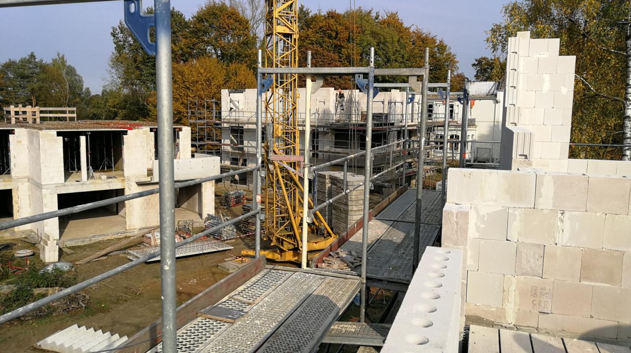 Wznoszenie ścian z białych bloczków na budowie domu, widoczne rusztowania, żuraw budowlany i częściowo wykończony budynek w tle.