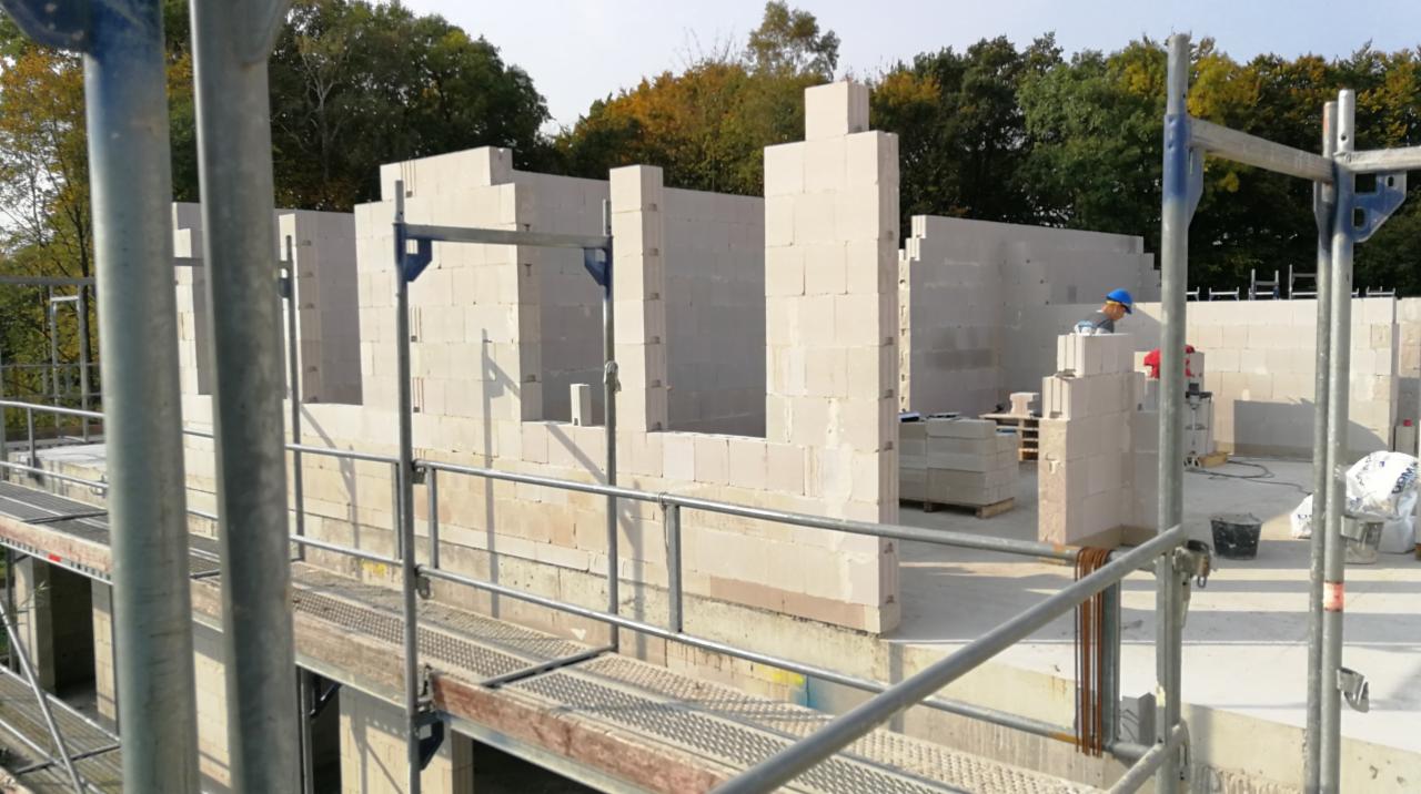 Wznoszenie ścian parteru z białych bloczków na placu budowy, widoczne rusztowanie i pracownik w niebieskim kasku.