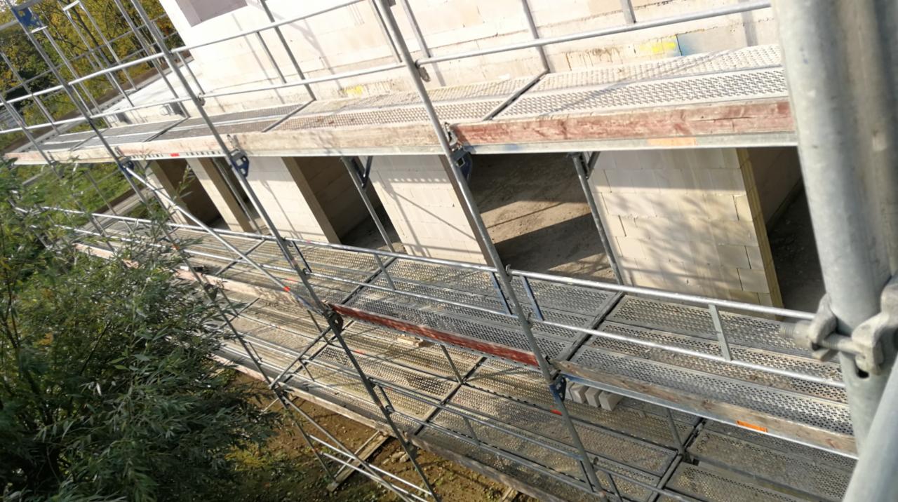 Wznoszenie ścian z bloczków na budowie domu, widok z rusztowania z perspektywy pracownika, fragment zieleni w tle.