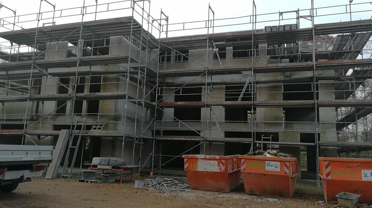 Wielopiętrowy budynek w trakcie budowy, otoczony rusztowaniami, z widocznymi pustakami betonowymi i pomarańczowymi kontenerami na gruz u dołu, częściowo zasłonięty przez zaparkowany samochód...