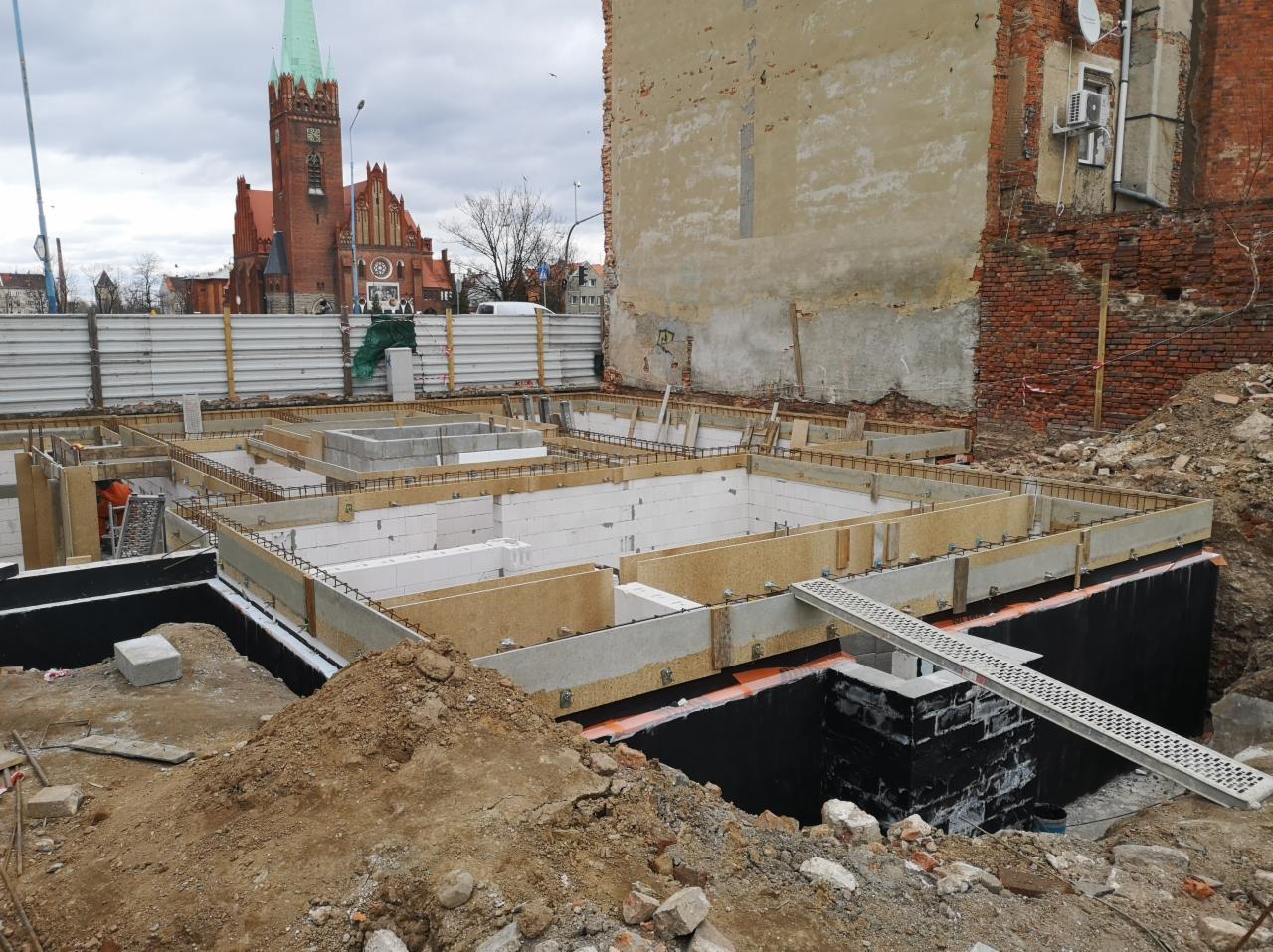 Fundamenty budynku w trakcie budowy, widoczne szalunki, zbrojenia, bloczki betonowe, izolacja przeciwwilgociowa oraz fragmenty ceglanego budynku w tle i kościół z zieloną wieżą w oddali.