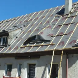 Dach dwuspadowy w trakcie krycia z widoczną membraną dachową z nadrukiem i drewnianą łatą kontrłatą, okna dachowe z folią zabezpieczającą, fragment elewacji budynku z deskowaniem pionowym i oknem.