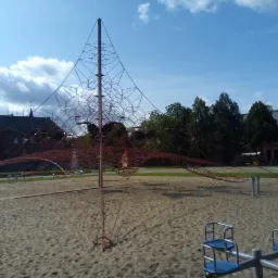 Konstrukcja wspinaczkowa z czerwonej liny na piaszczystym placu zabaw w słoneczny dzień, z widocznymi budynkami w tle.