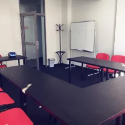 Jasna sala szkoleniowa z ciemnymi stołami w kształcie litery U, czerwonymi krzesłami, białą tablicą z kolorowymi magnesami, w tle wieszak na ubrania i otwarte szklane drzwi.