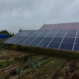 Instalacja paneli słonecznych na metalowej konstrukcji na polu, z widocznymi budynkami w tle pod pochmurnym niebem.