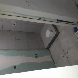 Szare płytki ceramiczne na ścianach i podłodze w trakcie montażu, widoczne krzyżyki dystansowe, ślady po szpachli na ścianie, przygotowanie do wykończenia łazienki.