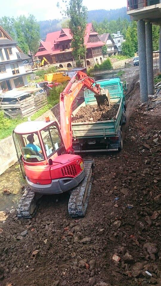 Czerwona minikoparka na gąsienicach nabiera ziemię do zielonej ciężarówki na placu budowy w górskim otoczeniu.