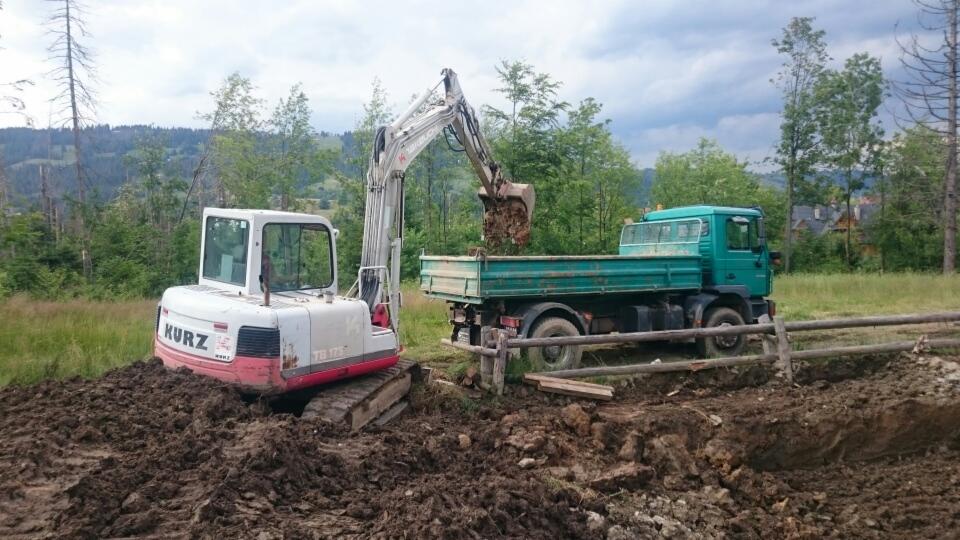 Biało-czerwona minikoparka marki Kurz na gąsienicach, z łyżką wysypującą urobek do zielonej ciężarówki na tle górskiego krajobrazu