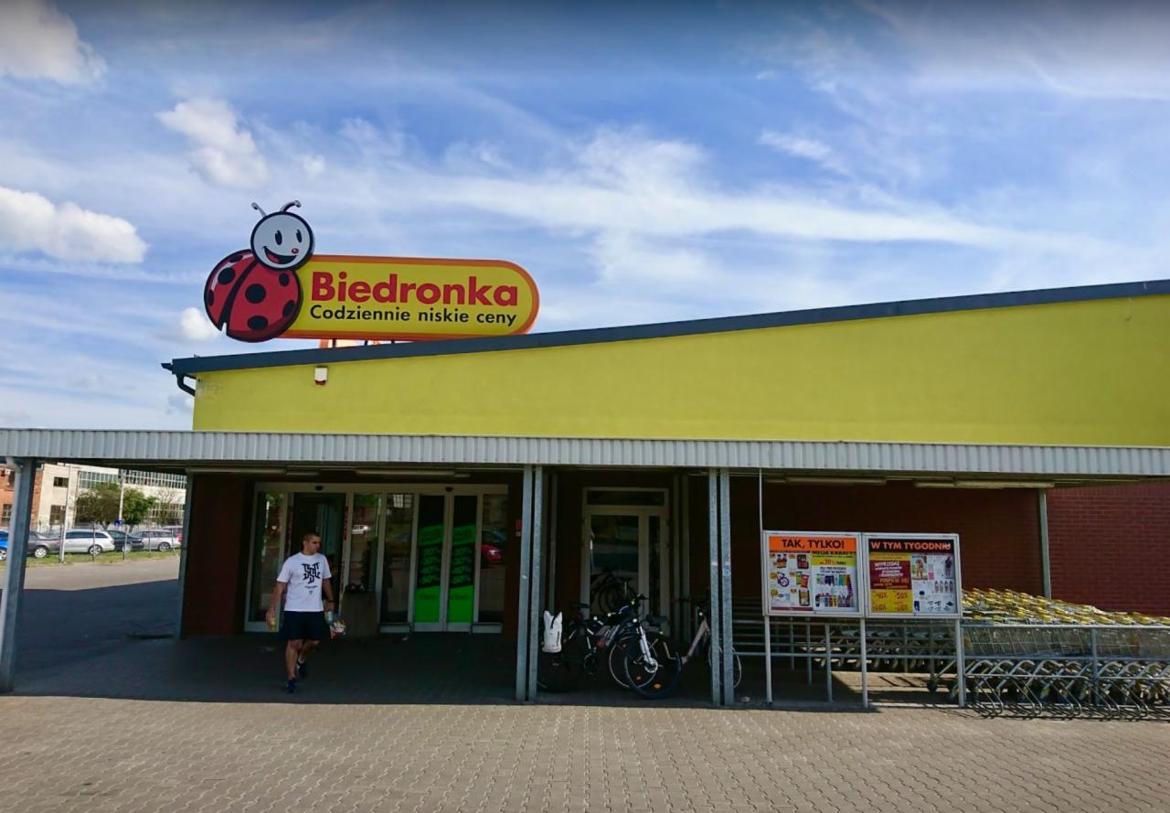 Fasada sklepu Biedronka z logo biedronki i napisem 'Codziennie niskie ceny', mężczyzna w białej koszulce wchodzi do sklepu, rowery zaparkowane przy wejściu, tablica z ofertami promocyjnymi z prawej...
