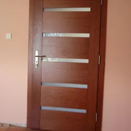 Ciemnobrązowe skrzydło drzwiowe z czterema poziomymi przeszkleniami, zamontowane w ościeżnicy w pokoju o ścianach w kolorze brzoskwiniowym i drewnianej podłodze.