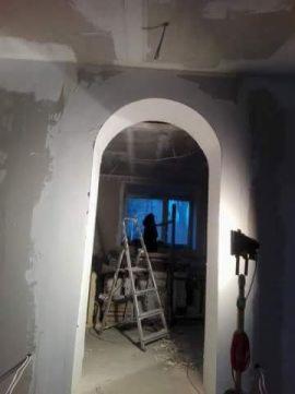 Łuk wejściowy w trakcie wykańczania, widoczne ślady po szpachlowaniu, drabina aluminiowa w tle, oświetlenie halogenowe na statywie, surowy stan ścian.