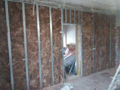 Ściana w trakcie izolacji termicznej, widoczna metalowa konstrukcja szkieletowa wypełniona brązową wełną mineralną, w tle fragment otworu drzwiowego z widocznymi materiałami budowlanymi.