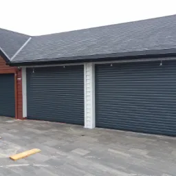 Trzy grafitowe bramy garażowe rolowane w budynku z drewnianą elewacją i szarym dachem, widoczne deski na brukowanym podjeździe.