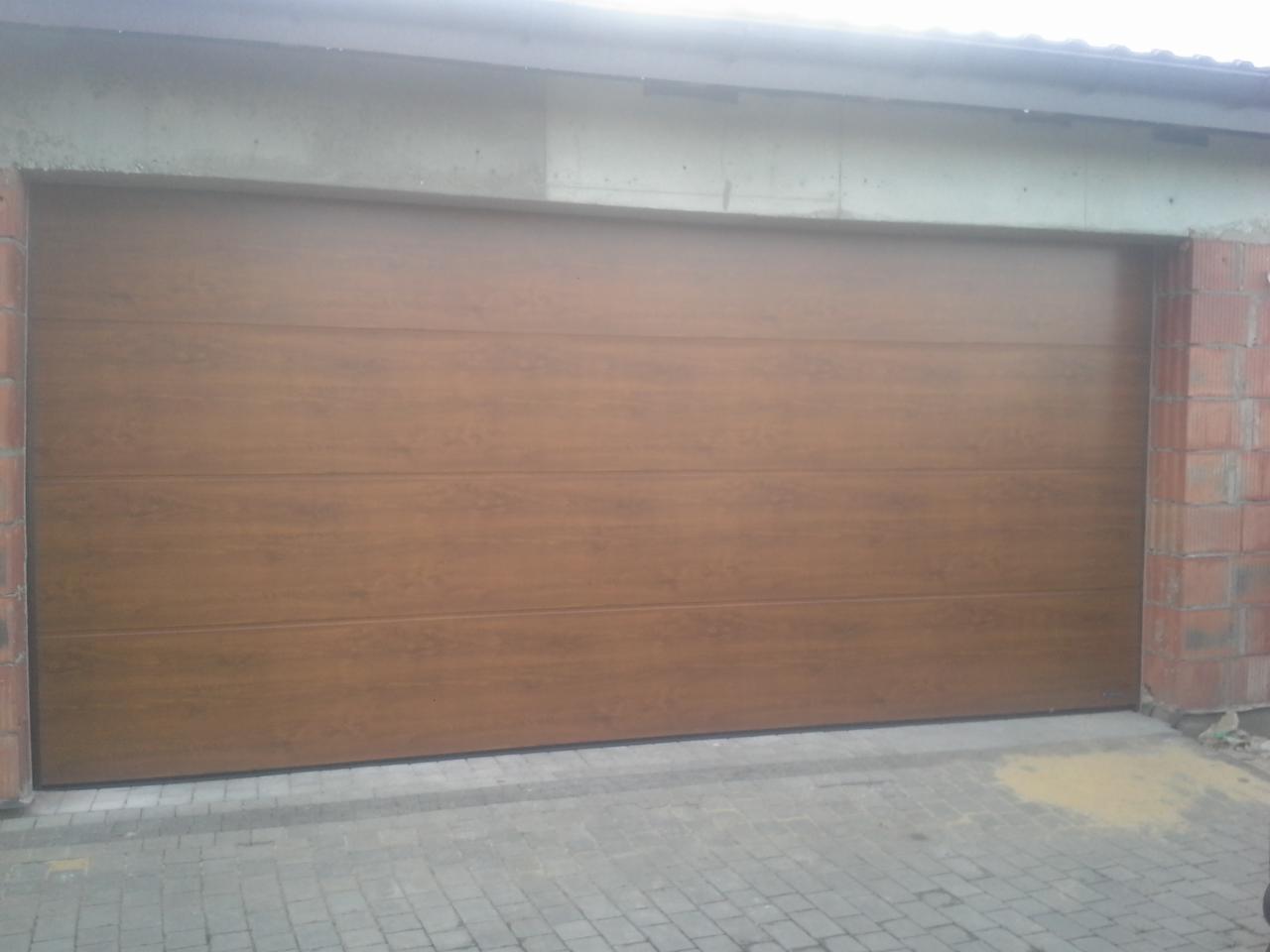 Zamknięta, segmentowa brama garażowa w kolorze drewna, z widocznym fragmentem muru z czerwonej cegły po prawej stronie, świeżo zamontowana, z widocznym cementem i kostką brukową przed bramą.