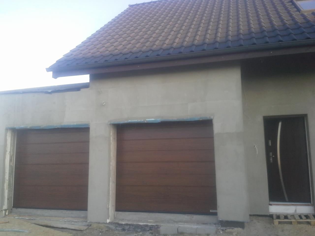 Dwa brązowe segmentowe w trakcie montażu w surowym stanie budynku, widoczna boczna elewacja z drzwiami wejściowymi.