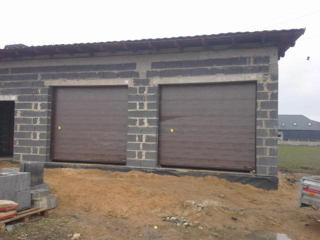 Dwie brązowe bramy garażowe segmentowe zamontowane w surowym budynku z bloczków betonowych, widoczne bloczki i piasek przed budynkiem.