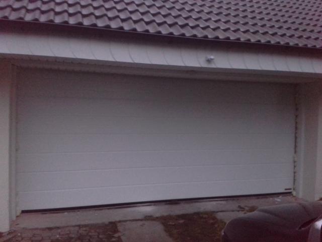 Zamknięta, segmentowa, biała brama garażowa z poziomymi przetłoczeniami, widoczna z zewnątrz budynku z ciemnym dachem.