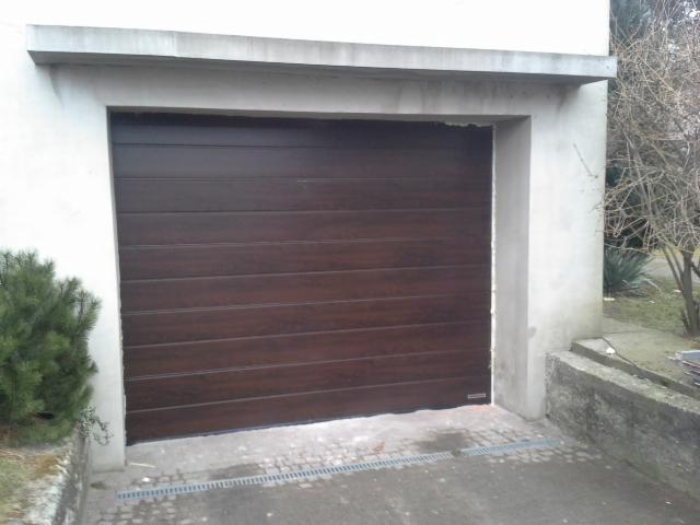 Ciemnobrązowa brama garażowa segmentowa z poziomymi panelami, zamontowana w surowym, betonowym otworze garażowym, widok z zewnątrz.