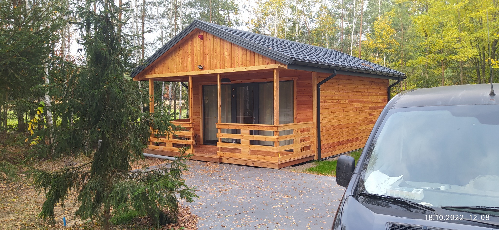 Nowo wybudowany, drewniany domek letniskowy z zadaszonym tarasem i czarnym dachem, widziany z perspektywy lekko z dołu, z fragmentem zaparkowanego samochodu w prawym dolnym rogu.