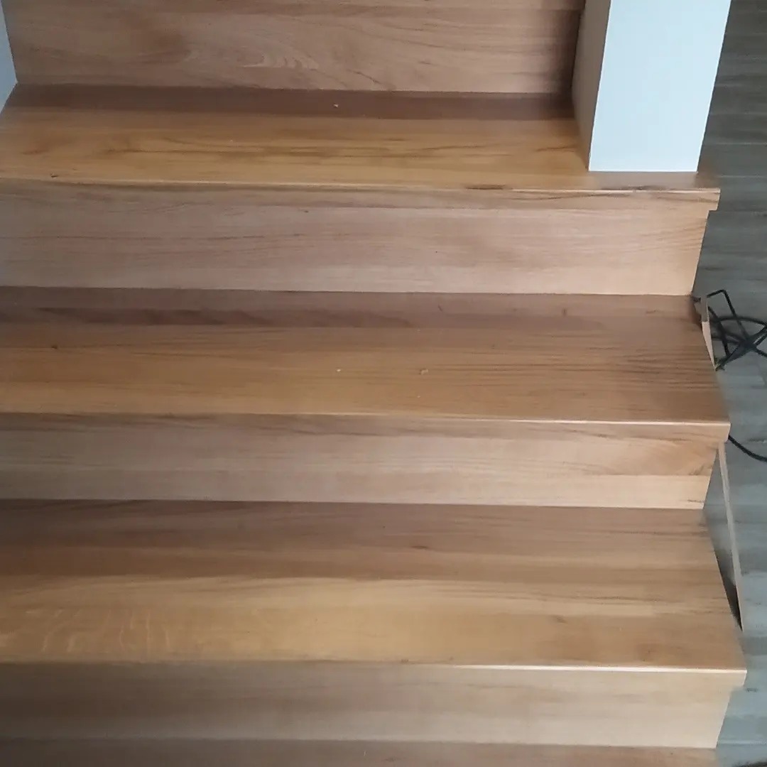 Drewniane schody z jasnego drewna, widoczne trzy stopnie, z fragmentem białej ściany po prawej stronie i kablem na podłodze.