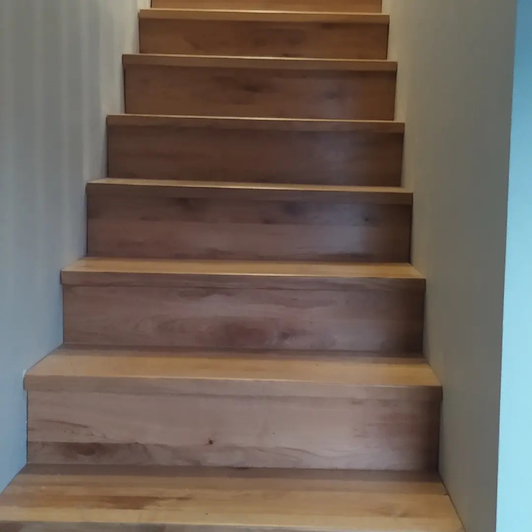 Drewniane schody z jasnego drewna, widok z dołu do góry, proste stopnie bez poręczy, ściana w kolorze złamanej bieli.