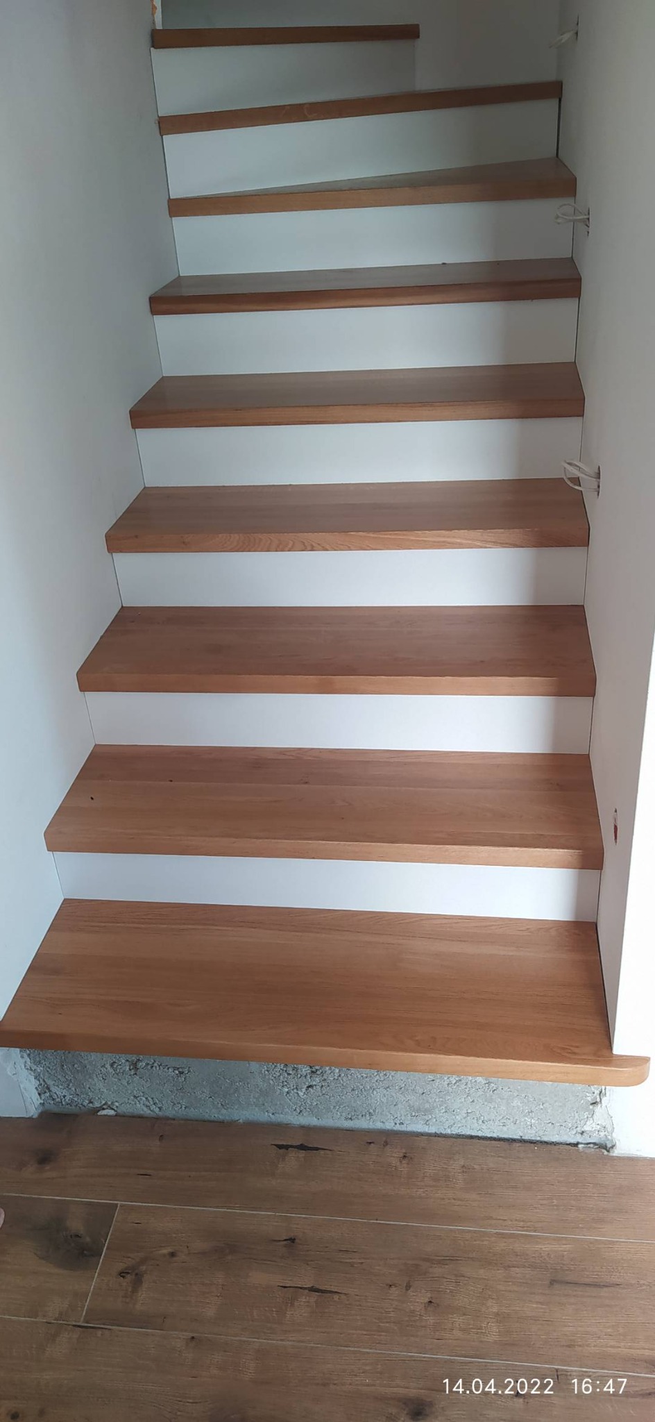 Schody drewniane z białymi podstopnicami, widok z dołu, z surowym wykończeniem podłoża i widoczną datą wykonania na podłodze.