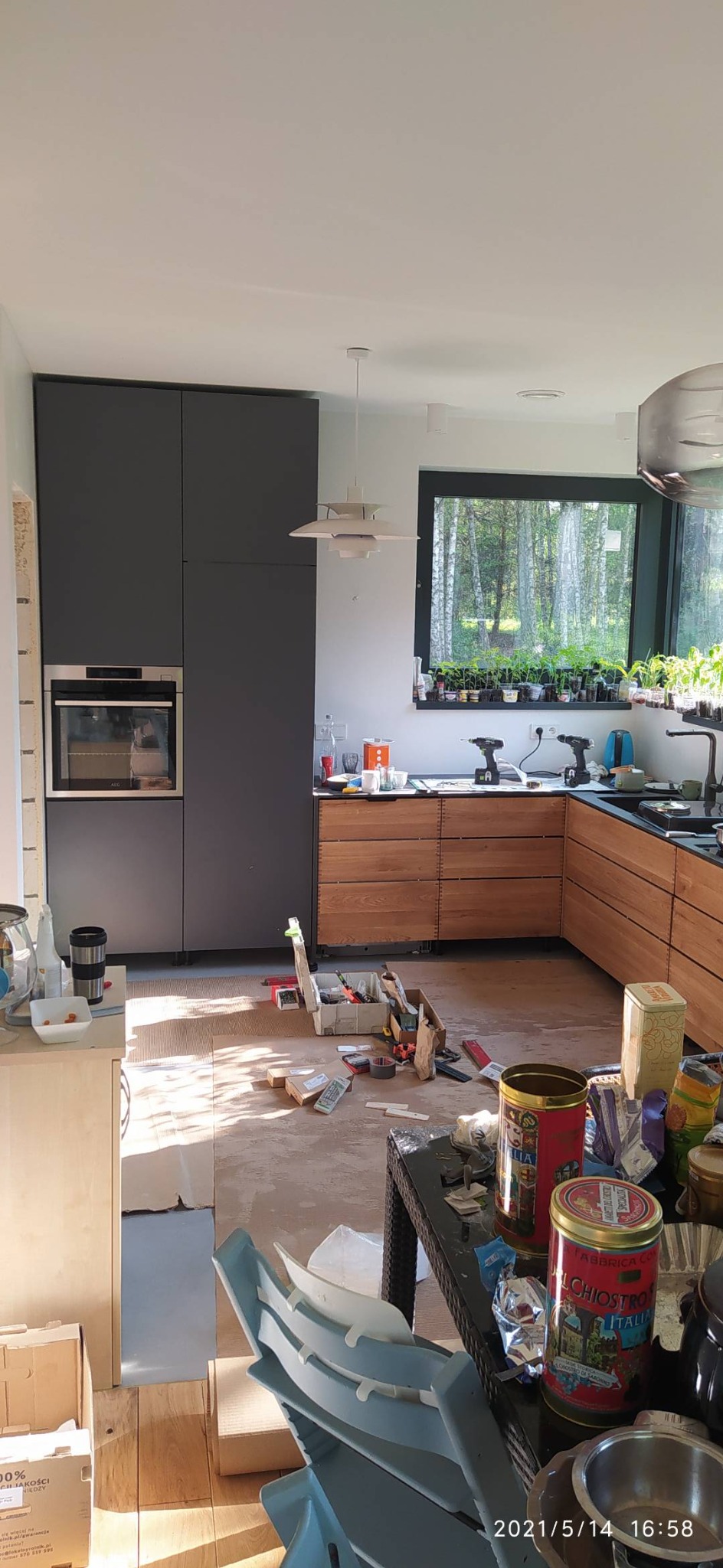 Kuchnia w trakcie remontu: widoczne szare szafki wiszące, drewniane szafki dolne, narzędzia i puszki leżące na podłodze oraz na stole, okno z widokiem na las.