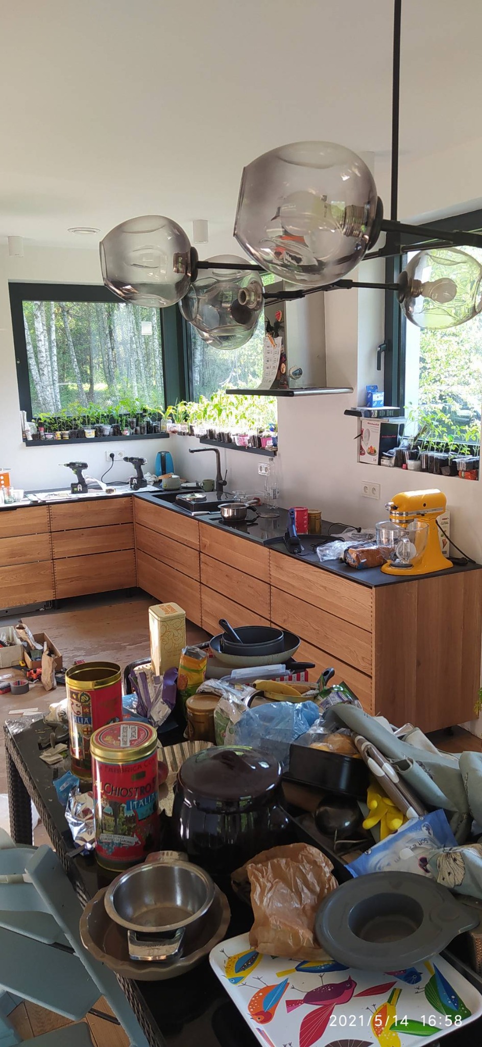 Kuchnia w trakcie prac wykończeniowych: widoczne drewniane szafki, czarny blat, okno z widokiem na las, narzędzia i akcesoria na blacie oraz stół z nieporządkiem.