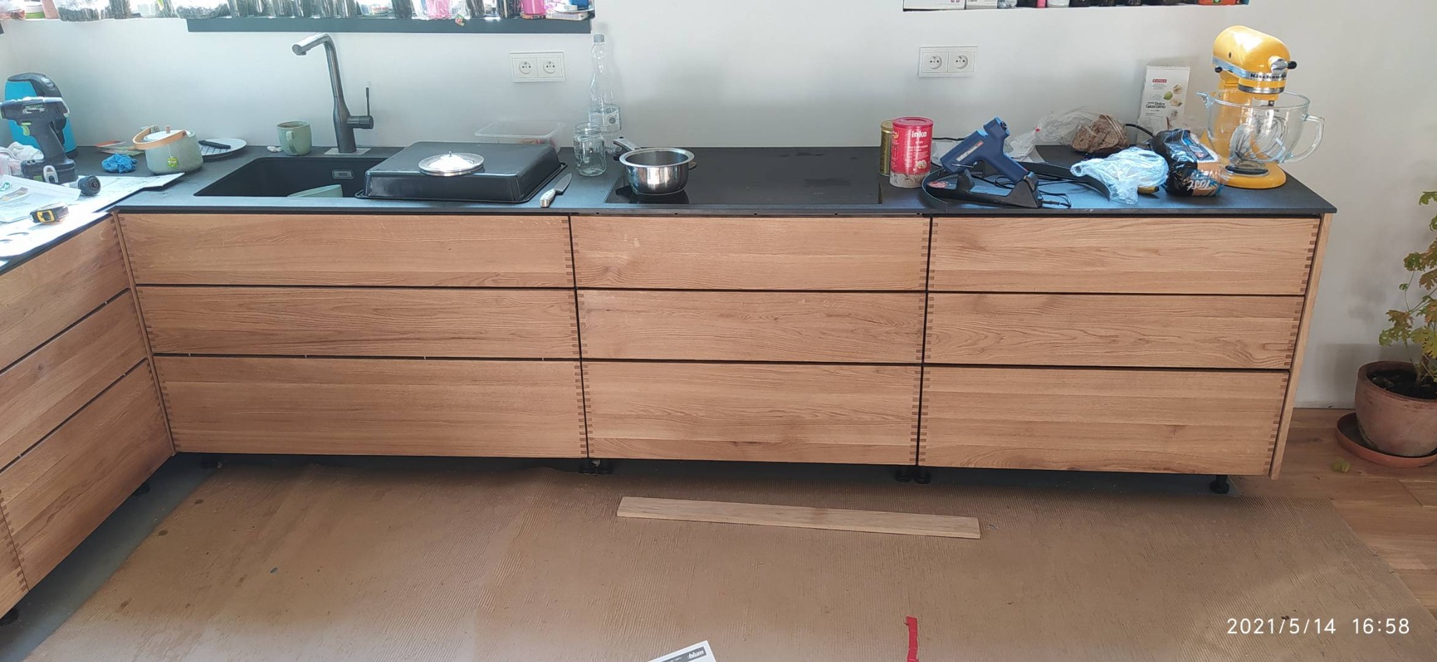 Nowoczesna kuchnia z drewnianymi frontami szuflad, czarnym blatem i sprzętami AGD na blacie, w trakcie prac wykończeniowych, widoczne narzędzia i akcesoria.