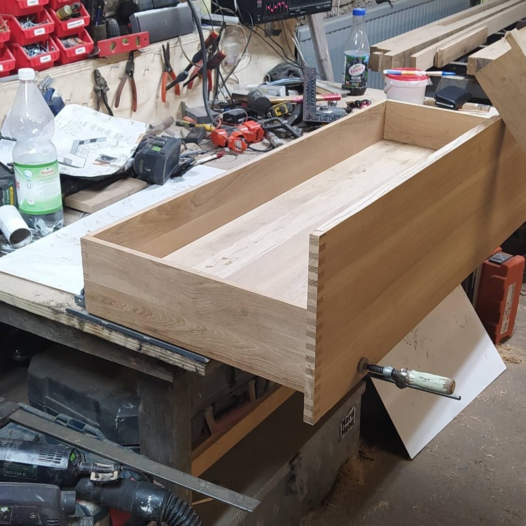 Surowy drewniany szuflada z widocznymi połączeniami na jaskółczy ogon, zamocowana ściskiem stolarskim, na warsztatowym stole z narzędziami w tle.