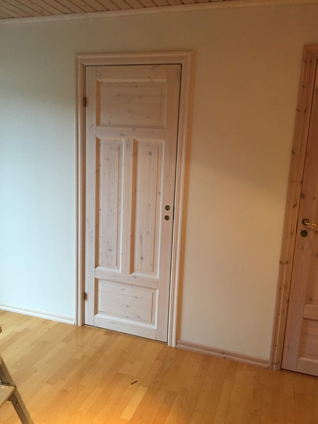 Jasne, surowe drewniane drzwi wewnętrzne z panelem, zamontowane w ścianie o kremowym odcieniu, na tle parkietu z widocznymi słojami drewna.