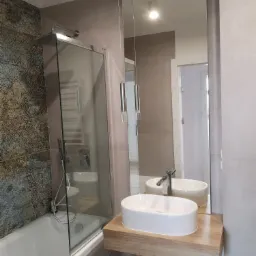 Nowoczesna łazienka z umywalką nablatową na drewnianym blacie, kabiną prysznicową ze szklaną ścianką i ozdobną płytką na ścianie przy wannie.