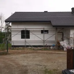 Nowo budowany dom jednorodzinny z ciemną dachówką, ocieplony styropianem, z widocznymi rusztowaniami i czarnymi ramami okiennymi, w trakcie prac wykończeniowych.