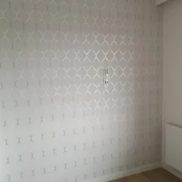 Ściana pomalowana na jasnoszaro z delikatnym, srebrzystym wzorem marokańskim, widoczne gniazdka elektryczne i fragment drewnianej podłogi.