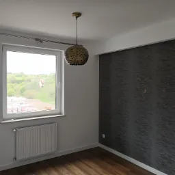 Pusty pokój po remoncie z drewnopodobną podłogą, szarą ścianą z fakturą, grzejnikiem pod oknem i ozdobną lampą wiszącą.